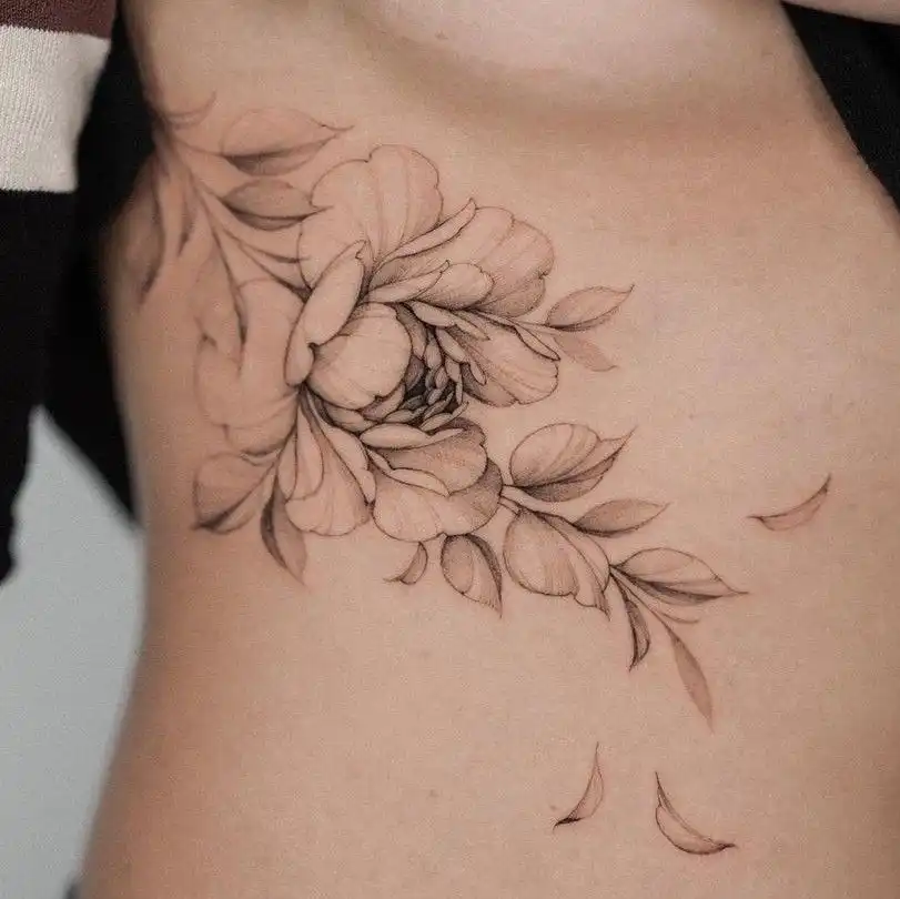 Delicate floral tattoo design on side of ribcage, featuring detailed petals and leaves in black and gray shading.