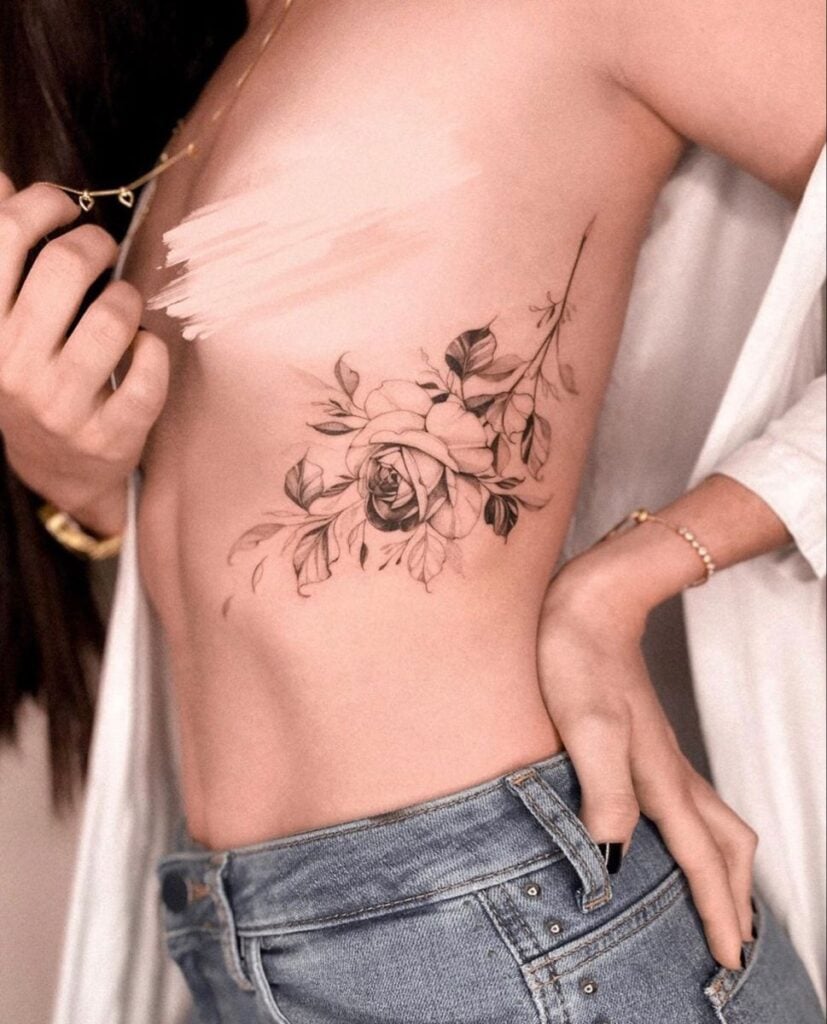 Elegant floral tattoo design on woman's side, featuring intricate roses and leaves. Jeans and delicate jewelry add a stylish touch.
