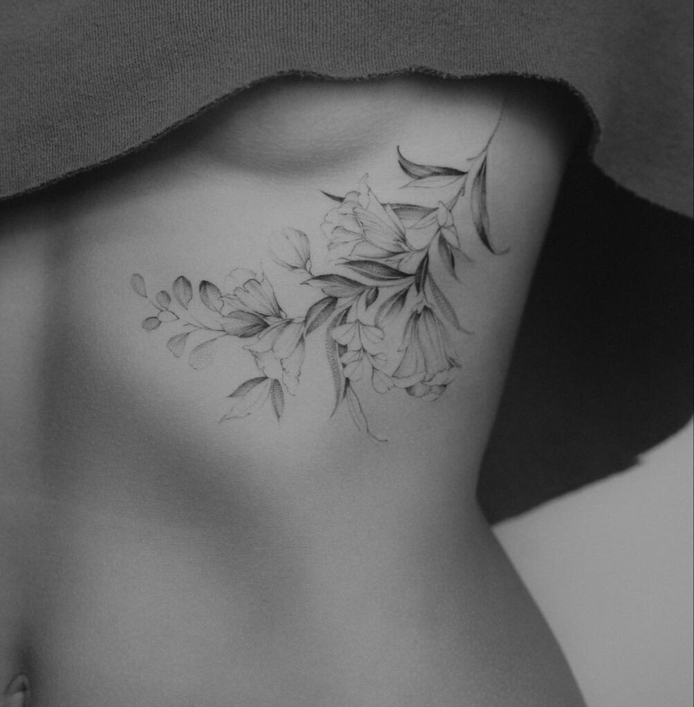 Black and white floral tattoo on a person's side under ribcage, showcasing detailed and delicate linework with leaves and flowers.