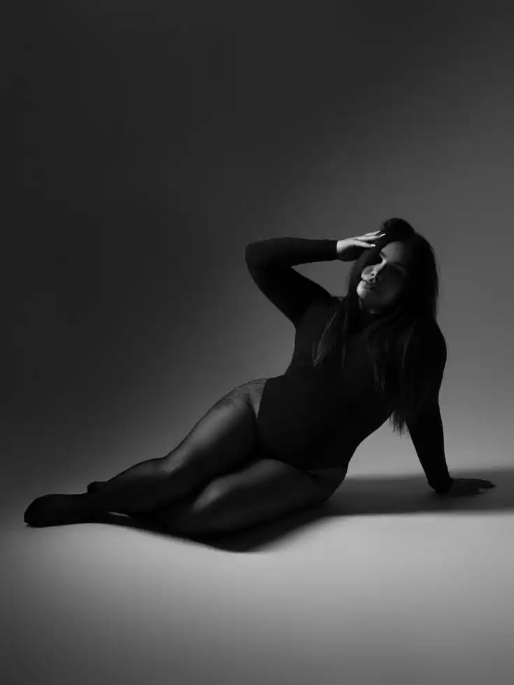 Black and white photo of a woman posing gracefully in a bodysuit, sitting on the floor with dramatic lighting and shadows.