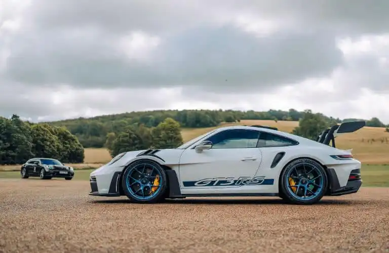 White sports car with GT3 RS decal parked on a scenic countryside road, featuring blue rims and a black spoiler under cloudy