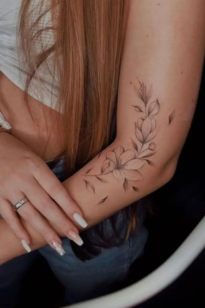 Elegant floral tattoo on forearm, complemented by long brown hair and manicured nails, showcasing artistic body ink style.