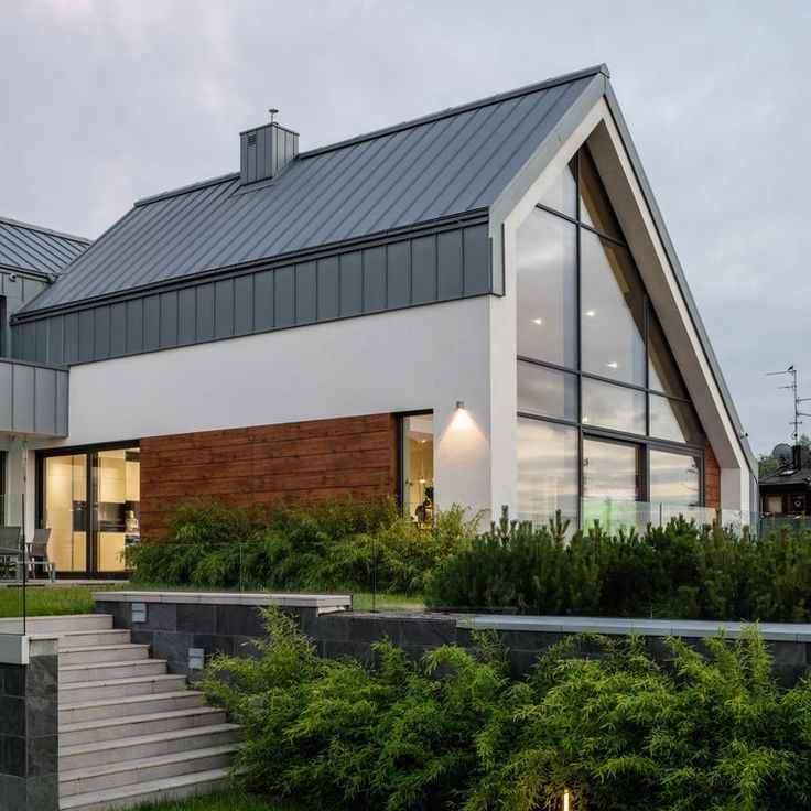 Modern house with large glass windows, metal roof, and wooden paneling, surrounded by greenery and a stone staircase in the