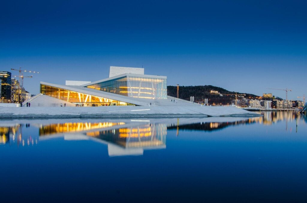 Modern architecture of Oslo Opera House reflects in calm water at dusk, showcasing its angular design under a clear blue sky.
