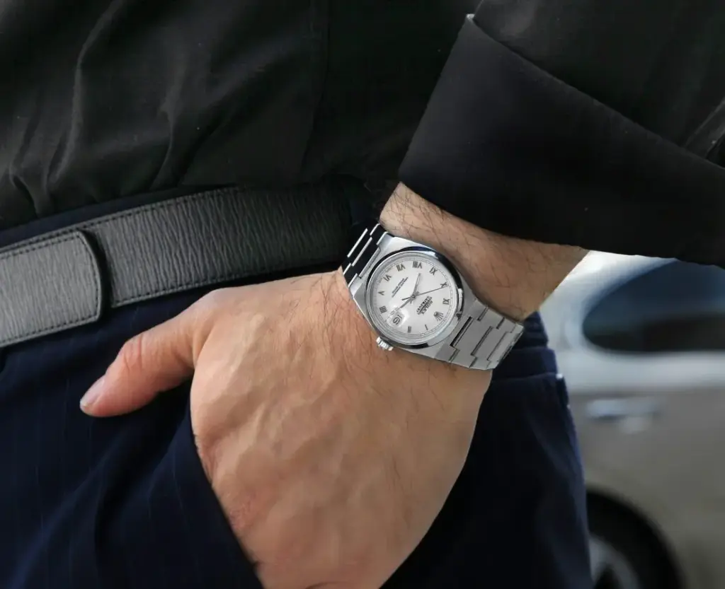 Man wearing a sleek, silver luxury wristwatch with a white dial and Roman numerals, hand in pocket. Fashionable style and elegance.