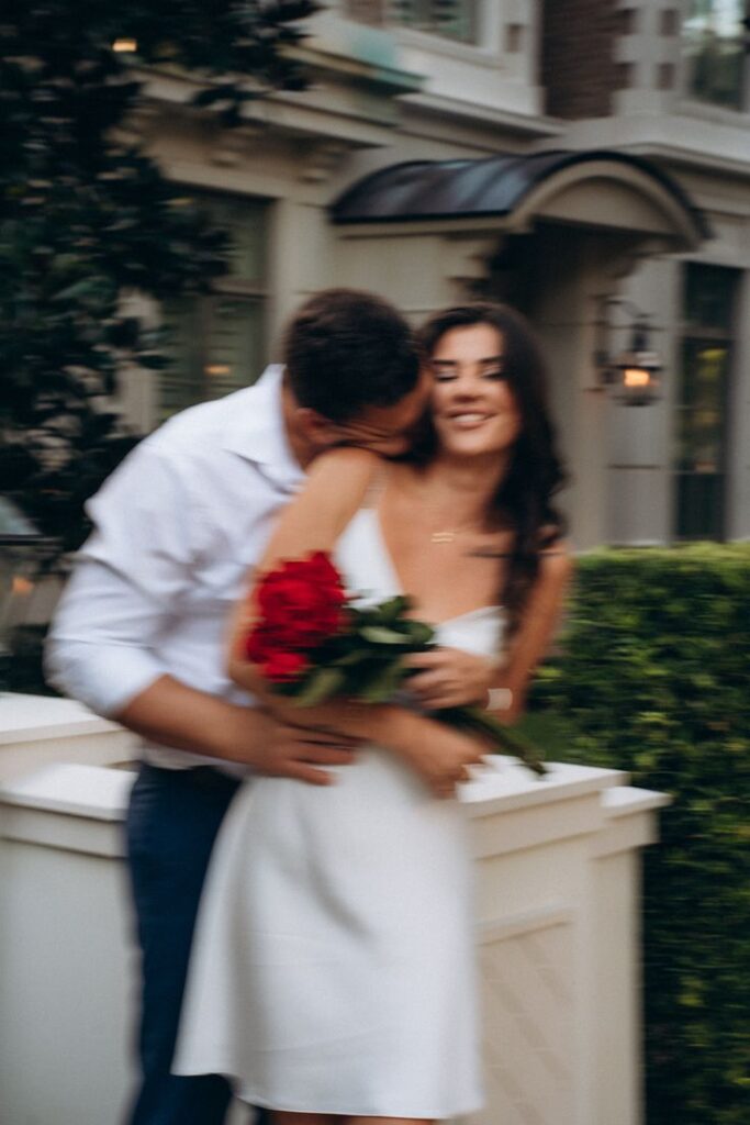 Romantic photoshoot _ Couples photography A couple embraces romantically outside, with the woman holding a bouquet of red roses, wearing a white dress, and smiling. | Sky Rye Design A couple embraces romantically outside, with the woman holding a bouquet of red roses, wearing a white dress, and smiling.