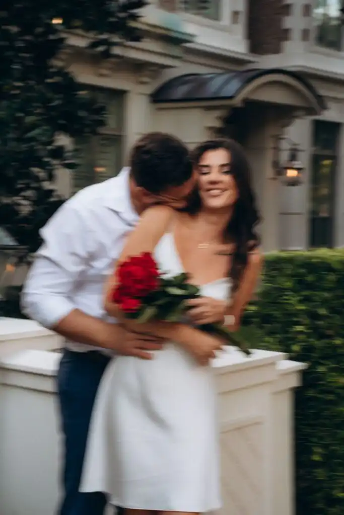 Romantic photoshoot _ Couples photography A couple embraces romantically outside, with the woman holding a bouquet of red roses, wearing a white dress, and smiling. | Sky Rye Design A couple embraces romantically outside, with the woman holding a bouquet of red roses, wearing a white dress, and smiling.