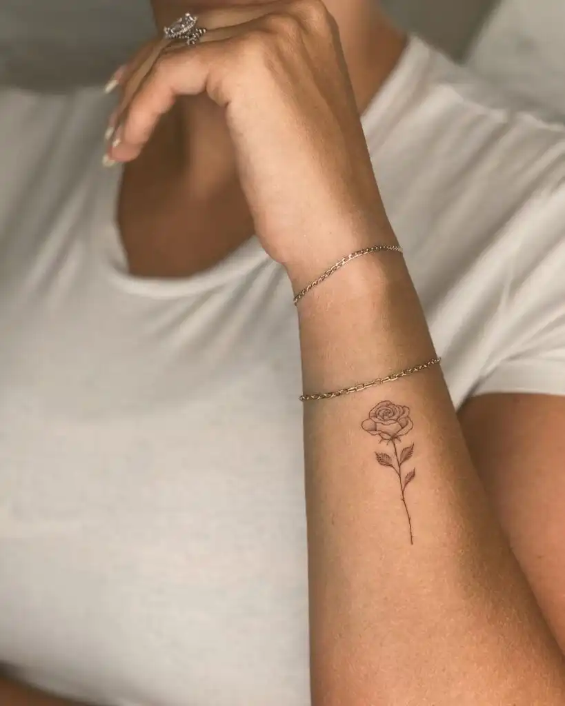 Minimalist rose tattoo on inner forearm, highlighted by a delicate chain bracelet and a white shirt backdrop.