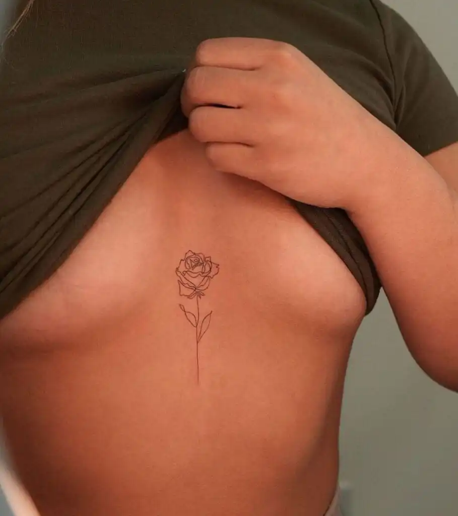 Minimalist rose tattoo on sternum, person holding up shirt to reveal art. Simple black line design emphasizes elegance and personal style.