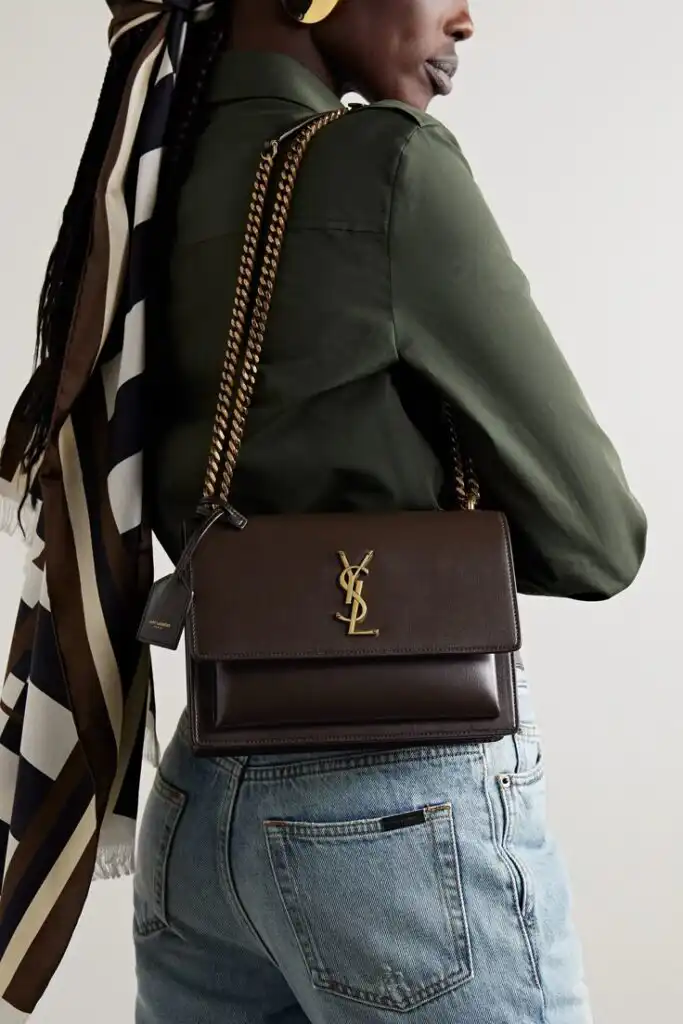 Woman in green shirt wearing designer YSL brown leather crossbody bag with gold chain strap and logo, paired with jeans and scarf.