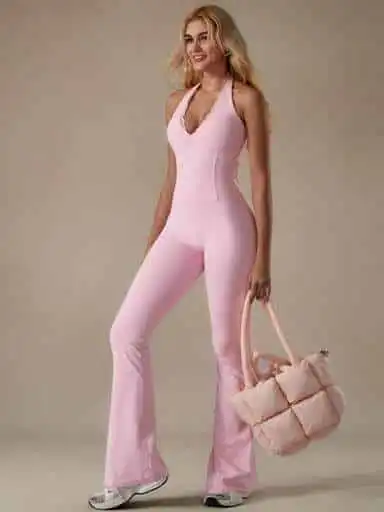 Smiling woman in a pink halter jumpsuit holding a matching quilted bag, standing against a beige background. Fashion and style concept.
