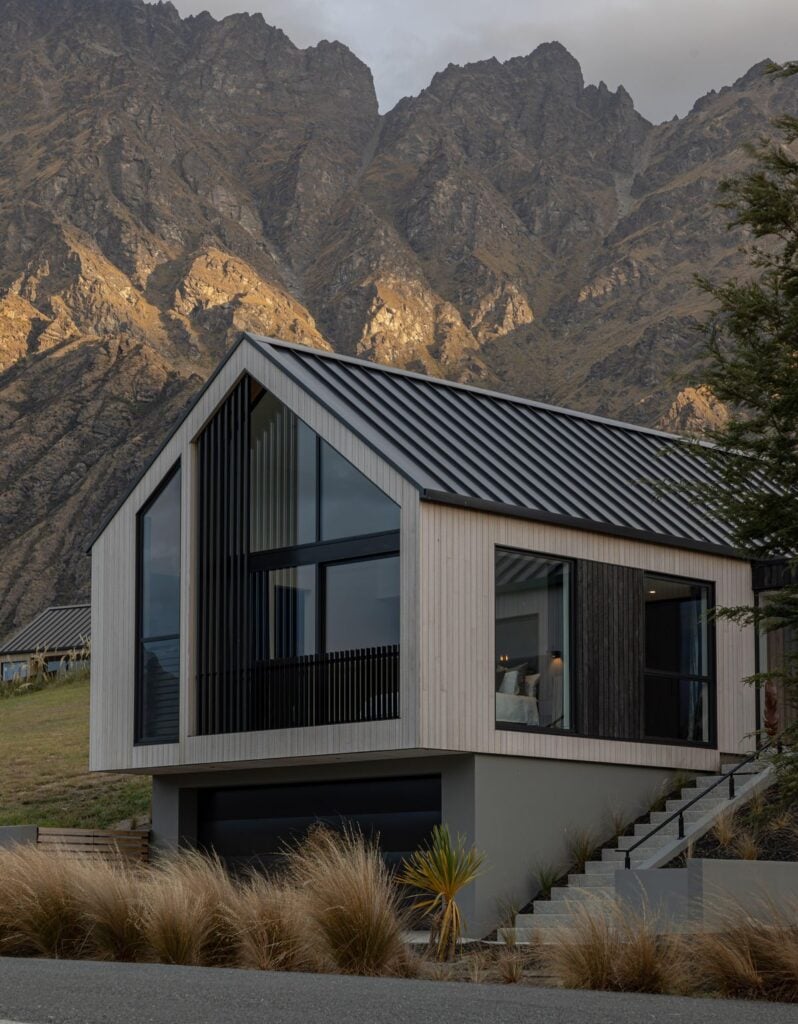 Modern mountainside house with sleek architecture, large windows, and a scenic backdrop of rugged cliffs, under a cloudy sky.