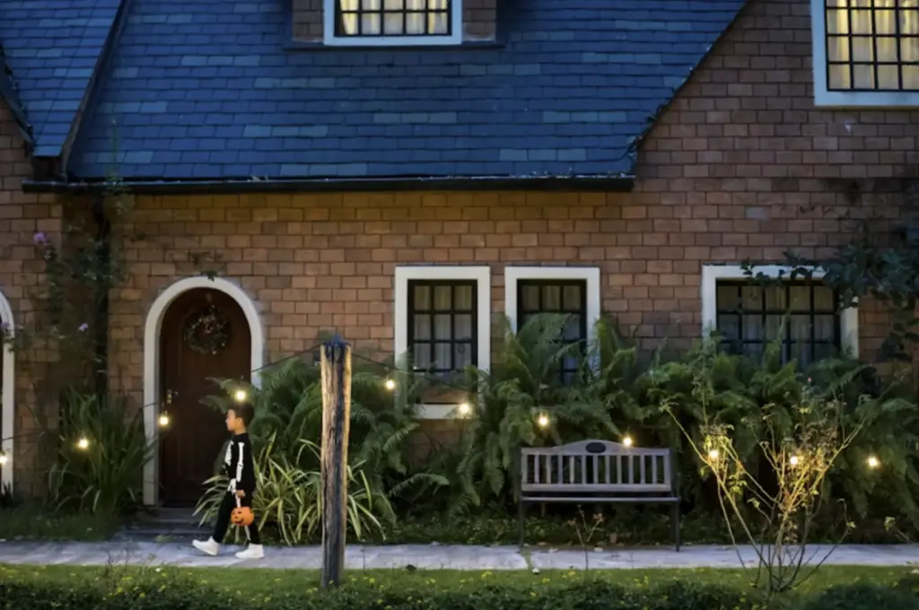 Child in skeleton costume trick-or-treating near a cozy, decorated house with soft string lights, during Halloween evening.
