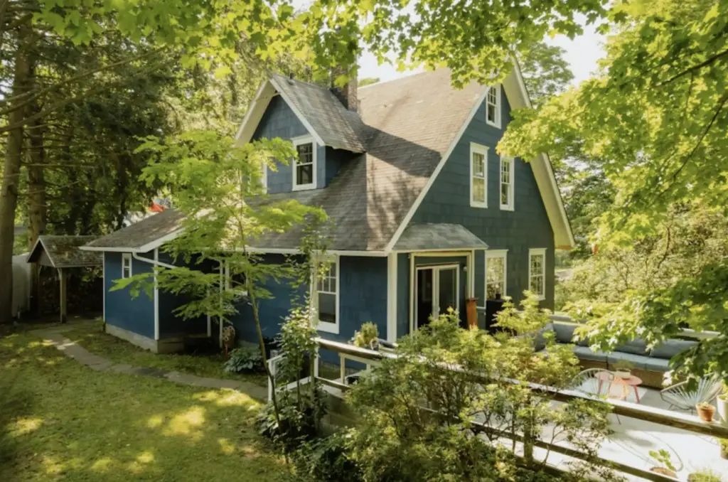 Charming blue cottage surrounded by lush trees, featuring a cozy patio with outdoor furniture, under a clear summer sky. Signs Of Quality Landscaping
