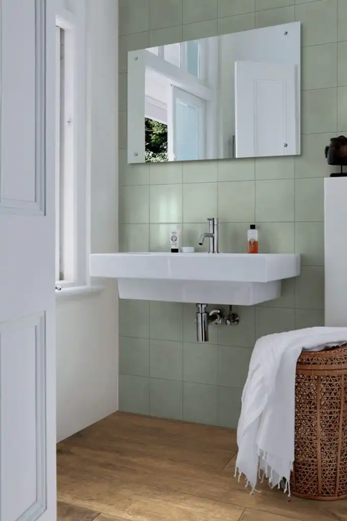 Small bathroom interior Modern bathroom with green tiled wall, floating sink, large mirror, and natural light. Cozy ambiance with wooden flooring and wicker basket. | Sky Rye Design Modern bathroom with green tiled wall, floating sink, large mirror, and natural light. Cozy ambiance with wooden flooring and wicker basket.