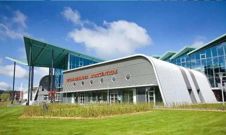 Modern sports center with unique architecture under a clear blue sky, surrounded by green lawn and glass windows reflecting sunlight.