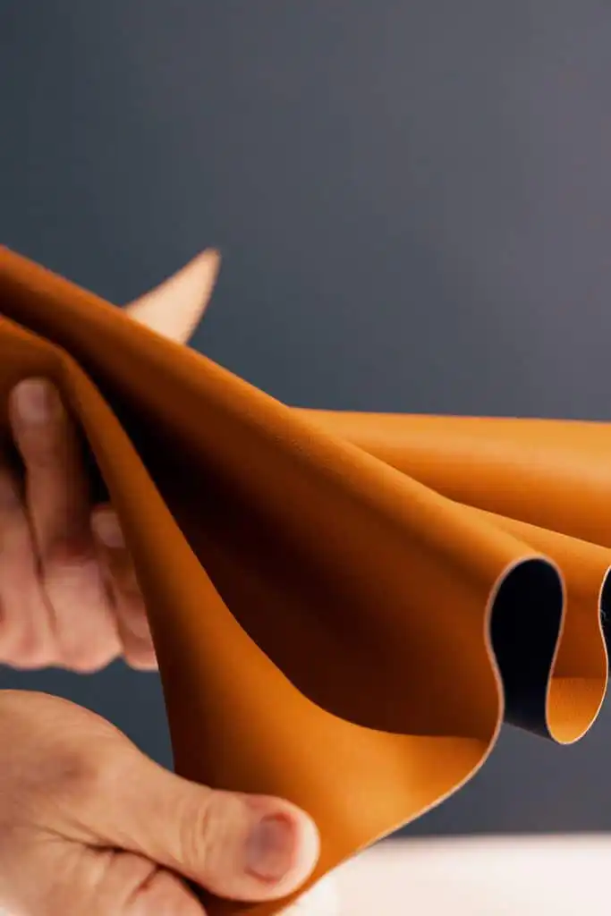 Person holding a piece of flexible, brown leather against a dark background, showcasing texture and quality for crafting or design.