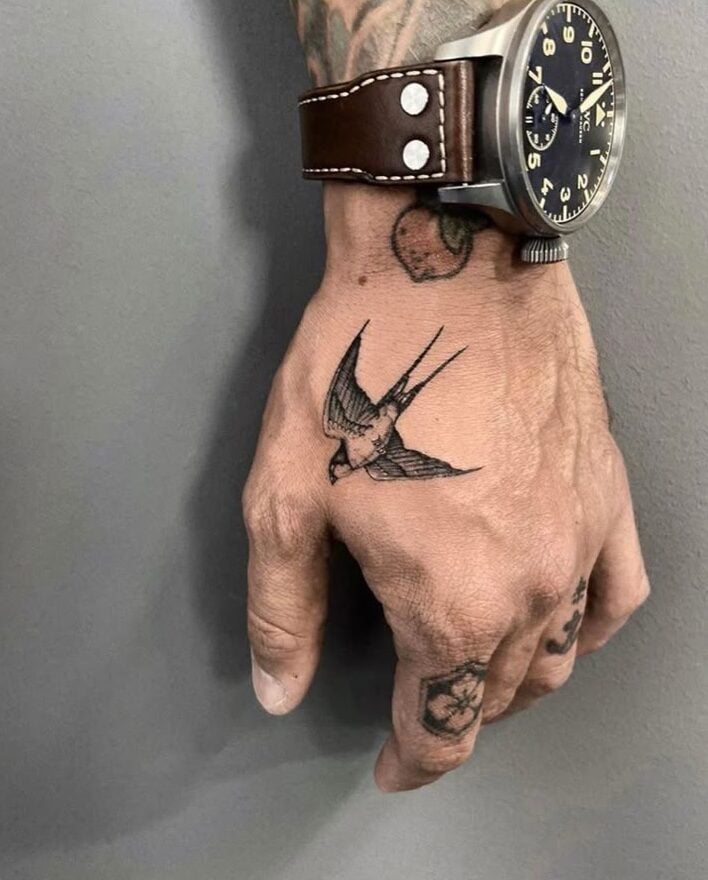 Hand with a flying swallow tattoo wearing a brown leather watch, against a gray background.