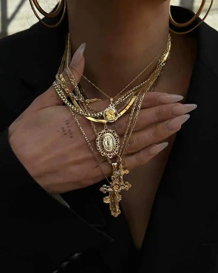 Stylish layered gold necklaces and hoop earrings, paired with elegant long nails on person in a black outfit.