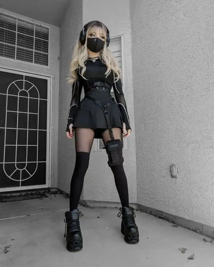 Young woman in edgy black outfit with mask, headphones, and platform boots stands confidently in front of a textured wall.