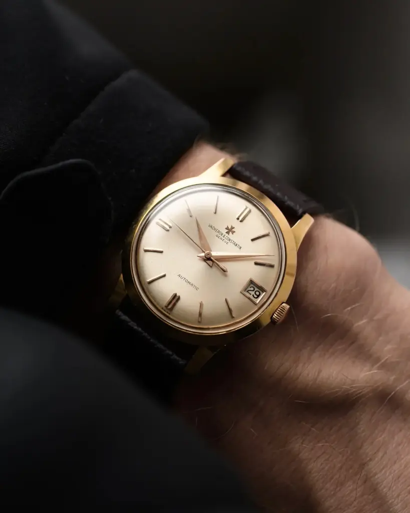 Close-up of a classic analog watch with a gold case, white dial, and leather strap on a wrist, showing the date at 3 o'clock position.