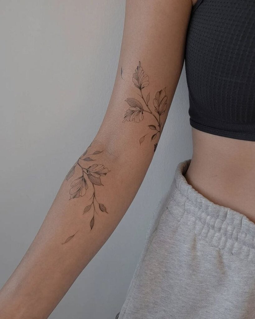 Minimalist floral tattoo on a person's forearm, wearing a black crop top and grey sweatpants, showcasing delicate and intricate design.