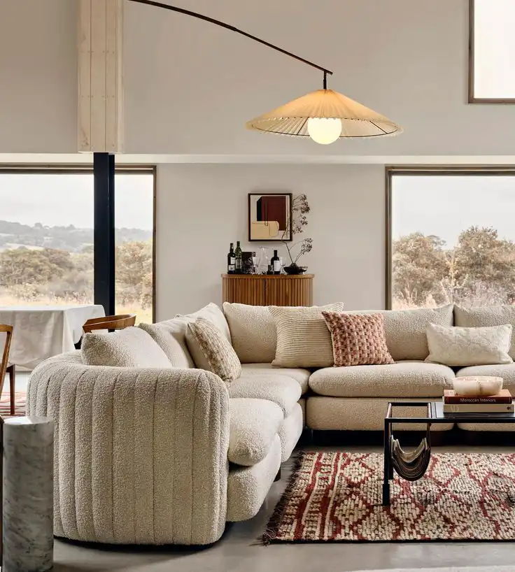 Modern living room with cozy beige sectional sofa, decorative pillows, patterned rug, and stylish pendant lamp, overlooking scenic outdoor view.