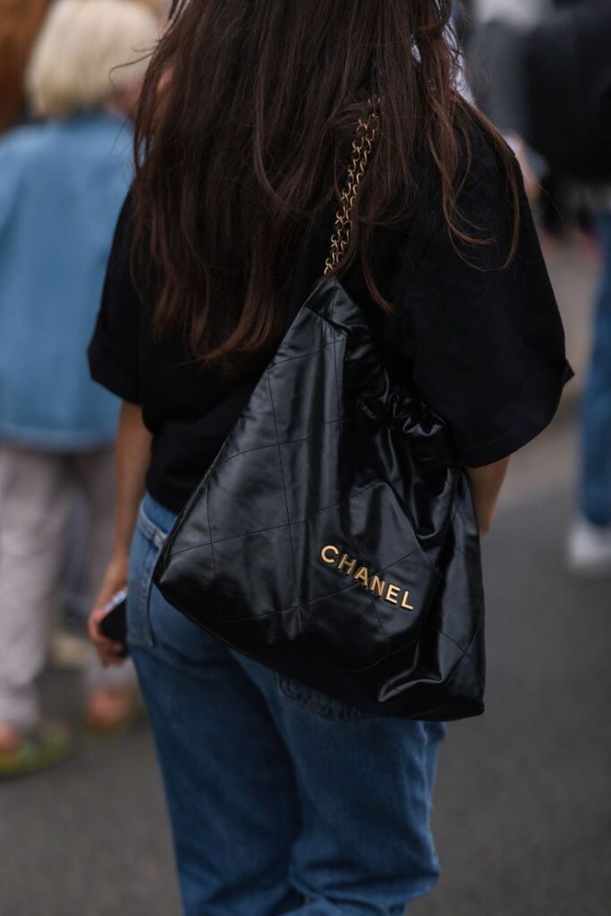 Vuelven los bolsos grandes y estos son los más bonitos Woman with long hair wearing black top and jeans carries a black quilted Chanel bag with gold chain strap outdoors. | Sky Rye Design Woman with long hair wearing black top and jeans carries a black quilted Chanel bag with gold chain strap outdoors.