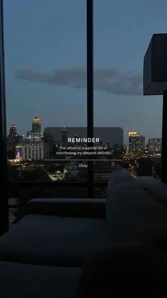 City skyline view at dusk through a window with a motivational reminder overlay about manifesting desires. Cozy couch in the foreground.