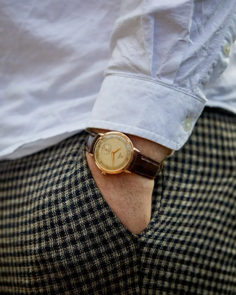 Close-up of a vintage watch with a brown leather strap on a person wearing a white shirt and checkered pants, hand in pocket.
