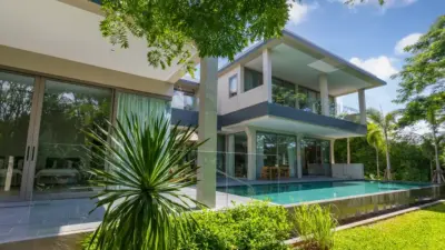 Modern two-story house with large windows, surrounded by lush greenery, featuring a sleek patio and inviting swimming pool.