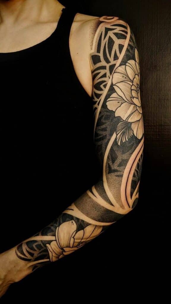 a2c863f0-43b2-418d-bac1-fd17e5c1cb16 Intricate arm sleeve tattoo featuring floral and geometric designs in black and beige tones on a person wearing a black top. | Sky Rye Design Intricate arm sleeve tattoo featuring floral and geometric designs in black and beige tones on a person wearing a black top.
