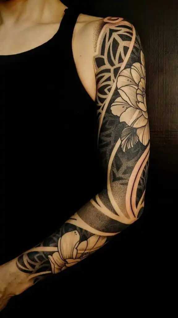 Intricate arm sleeve tattoo featuring floral and geometric designs in black and beige tones on a person wearing a black top.