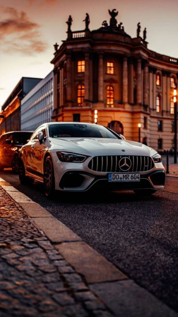 Sleek white Mercedes-Benz car parked on cobblestone street at sunset, historic building in background. Luxurious urban