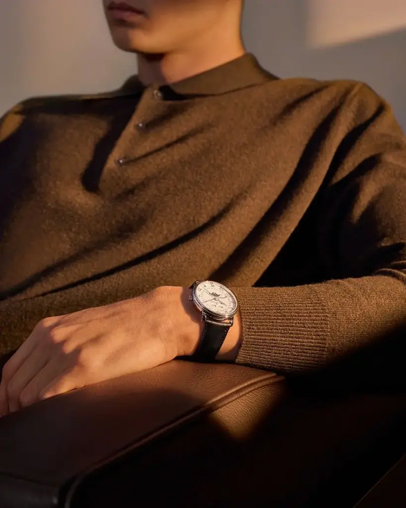 Man in brown sweater showing elegant wristwatch with leather band, seated in sunlight. Perfect for style and luxury fashion inspiration.