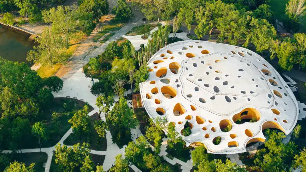 Aerial view of a unique architectural building with numerous circular openings, surrounded by lush green trees and winding