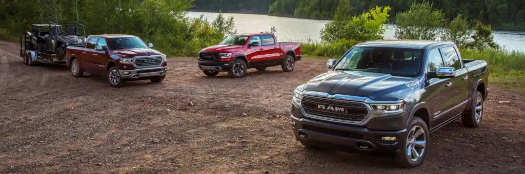 Three Ram trucks on a scenic dirt road by a lake, showcasing towing capability and rugged design.