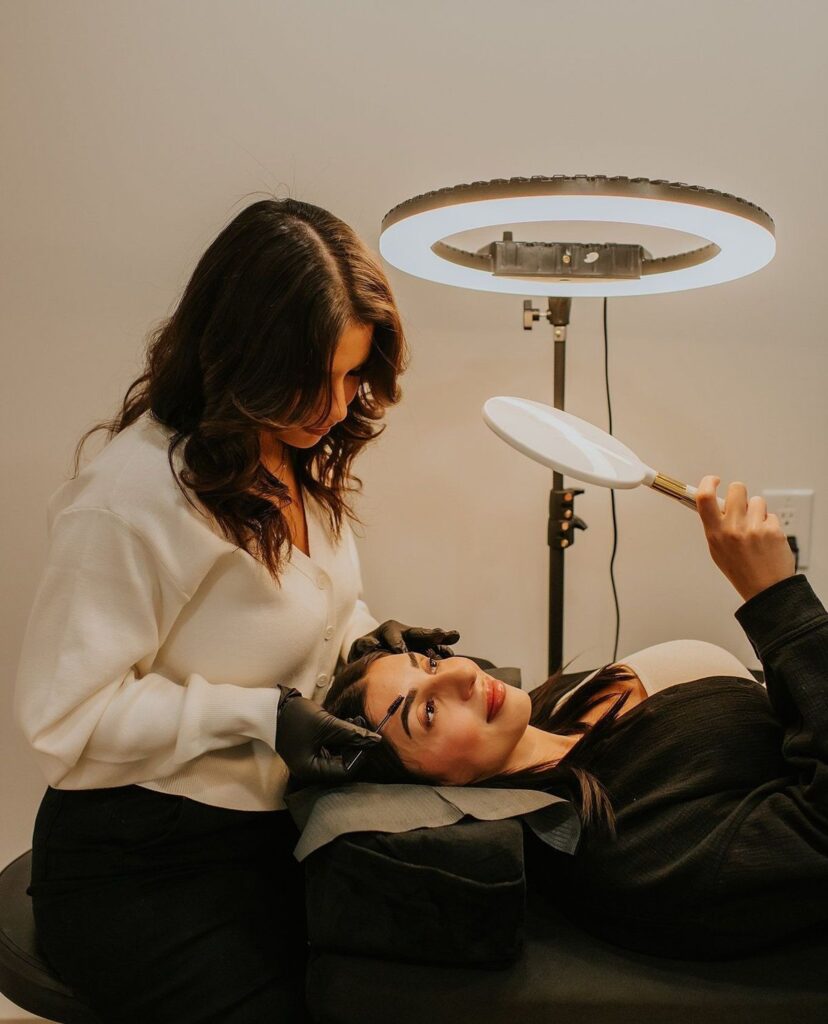 b9931603308fb695fae3f8d7f9a6757e Woman receiving eyebrow treatment in a beauty salon under a ring light, with an aesthetician applying makeup while she holds a mirror. | Sky Rye Design Woman receiving eyebrow treatment in a beauty salon under a ring light, with an aesthetician applying makeup while she holds a mirror.