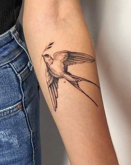 Forearm tattoo of a realistic flying bird with an olive branch, symbolizing peace and freedom, against a denim background.