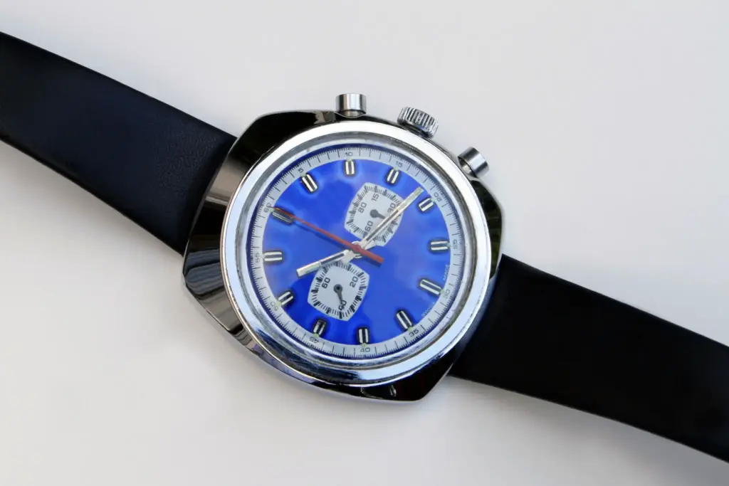 Vintage chronograph wristwatch with a blue face, silver case, and black leather strap on a white background.