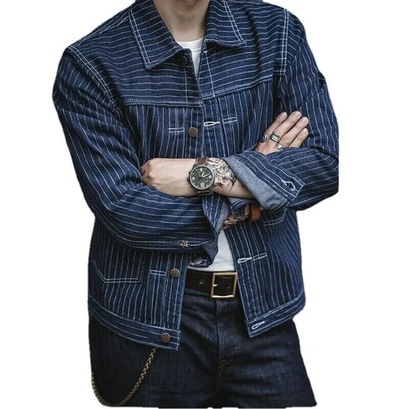 Man in a striped denim jacket and jeans with folded arms, showcasing a watch and rings against a white background.