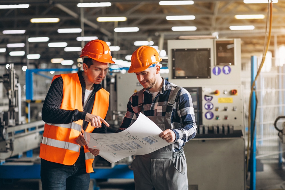 building-services-engineer Two engineers in safety gear discussing blueprints in a modern factory setting. Industrial machinery visible in the background. | Sky Rye Design Two engineers in safety gear discussing blueprints in a modern factory setting. Industrial machinery visible in the background.
