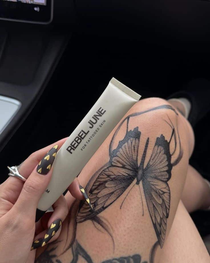 butterfly tattoo care Tattooed leg featuring a butterfly design with a hand holding Rebel June skincare tube, highlighting tattoo care. | Sky Rye Design Tattooed leg featuring a butterfly design with a hand holding Rebel June skincare tube, highlighting tattoo care.