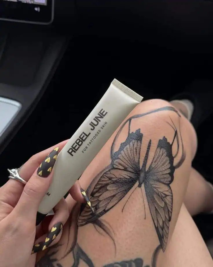 Tattooed leg featuring a butterfly design with a hand holding Rebel June skincare tube, highlighting tattoo care.