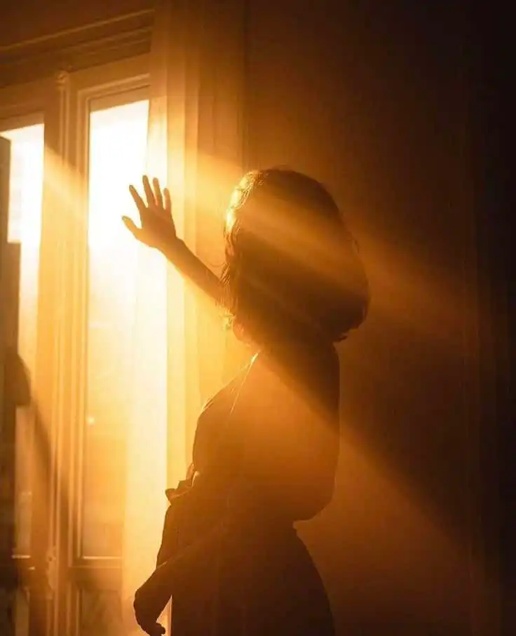 Silhouette of a woman in sunlight, reaching towards a window with golden sun rays illuminating the room. Warm and serene atmosphere.