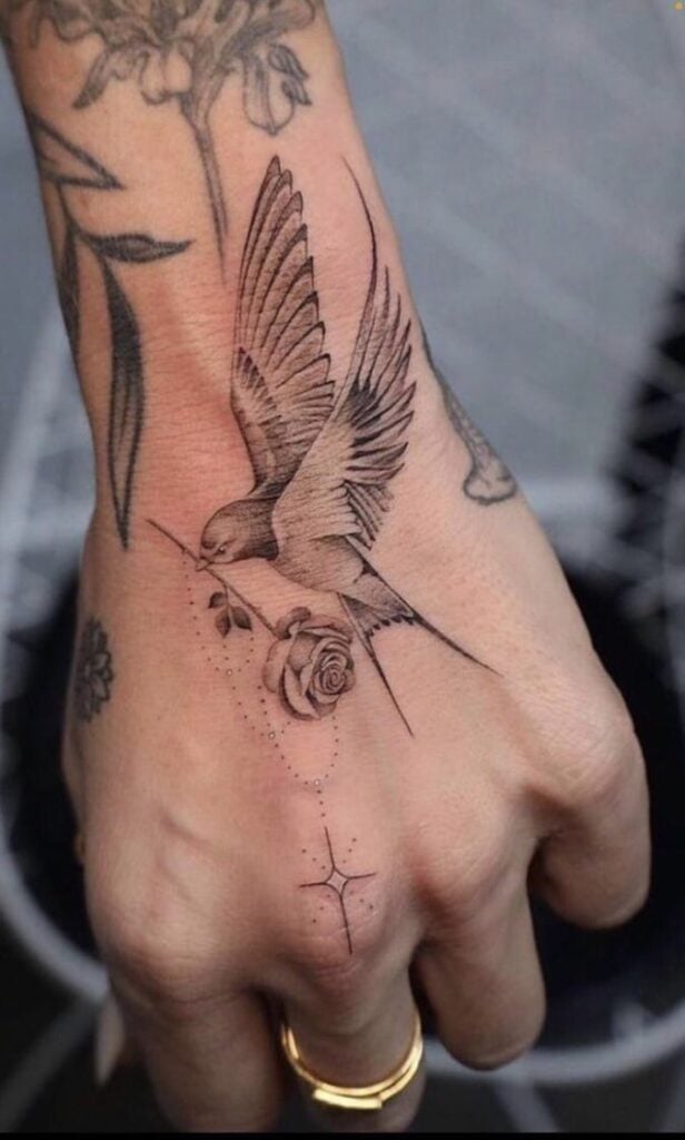 Hand tattoo of a bird carrying a rose, symbolizing freedom and beauty, with delicate linework and star accents on the skin.