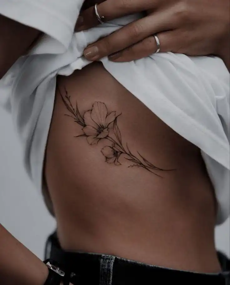 Delicate floral tattoo on side torso, showcasing elegant flowers and leaves under white shirt, highlighting minimalist body art.