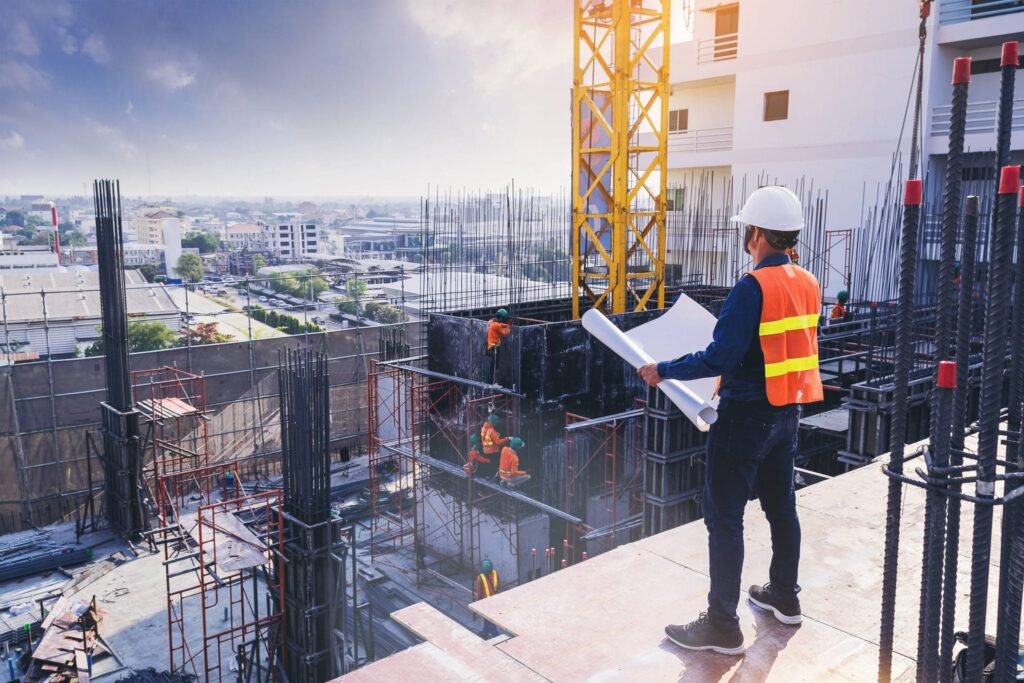 civil-eng-undergrad-banner Construction site with engineer in hard hat and safety vest, holding blueprints, overseeing workers and structures under construction. | Sky Rye Design Construction site with engineer in hard hat and safety vest, holding blueprints, overseeing workers and structures under construction.
