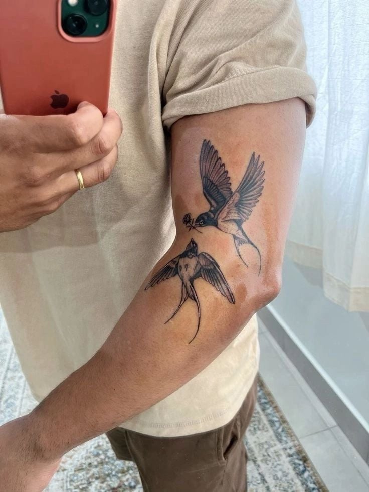 Arm tattoo of two flying swallows on a man taking a mirror selfie with a smartphone. Light brown shirt rolled up.