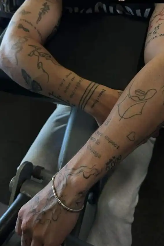 Tattooed arms rest on gym equipment, with intricate designs and text art clearly visible, conveying a blend of fitness and personal style.